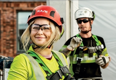 Två byggarbetare på en arbetsplats bär säkerhetsutrustning. I förgrunden syns en leende kvinna med en röd skyddshjälm märkt "KASK", skyddsglasögon och en neongrön arbetsjacka med reflexer. Hon har säkerhetsselar på sig. Bakom henne står en man i en vit skyddshjälm, solglasögon och en svart-grön arbetsjacka med reflexer. Bakgrunden visar en byggarbetsplats med en tegelbyggnad och en plasttäckning.