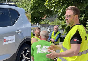 Bil med text Byggnads på väg samt man i varselväst pratar till andra män.