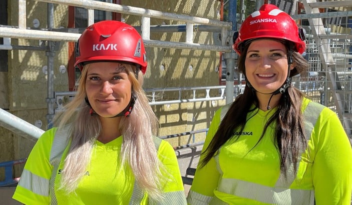 Två kvinnliga byggnadsarbetare med röda hjälmar och varseltröjor.