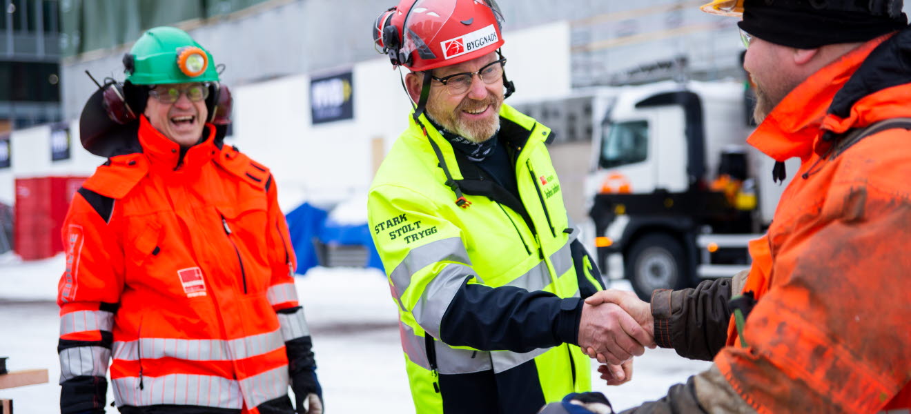 Johan Lindholm hälsar på byggnadsarbetare samtidigt som en annan byggnadsarbetare står bakom och ser väldigt glad ut..